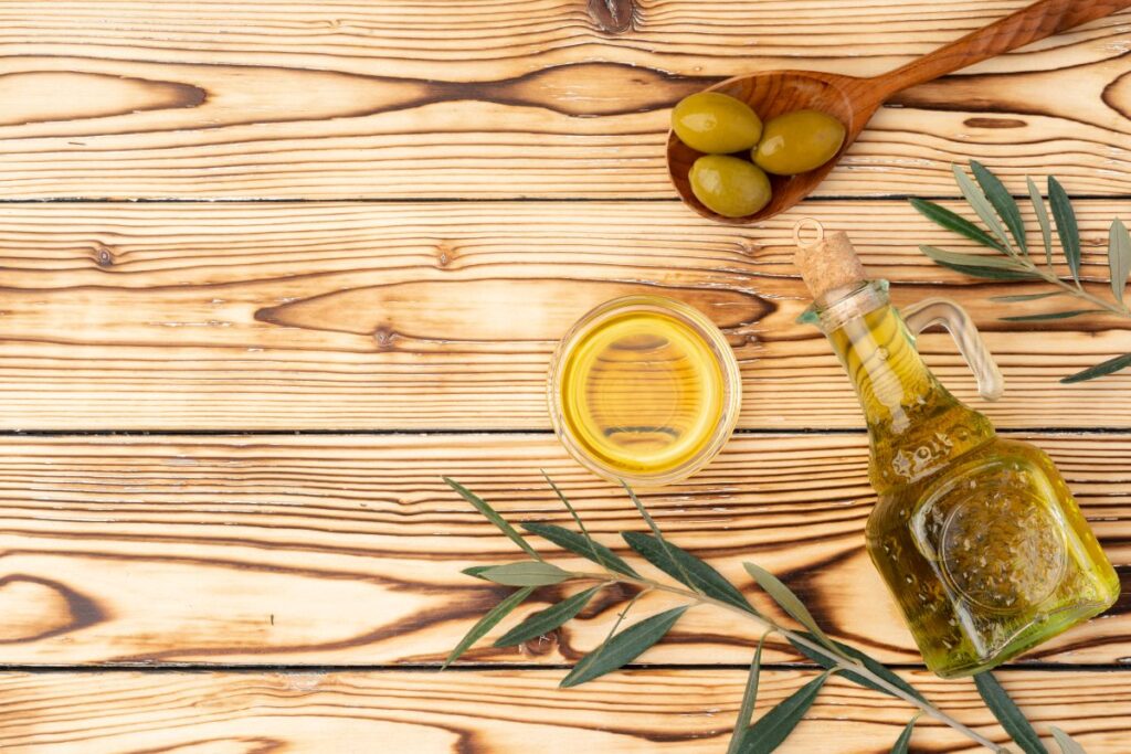 Un bocal en verre d'hiuile d'olive, un bol en verre avec de l'huile d'olive et des olives vertes dans une cuillères en bois le tout sur une table en bois
